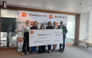 Five people stand smiling indoors, holding an oversized $300,000 check from GSK Canada, awarded to the School of Pharmacy at University of Waterloo for their immunization initiative. University and GSK logos are displayed behind them.
